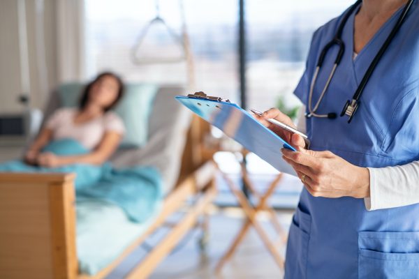 Unrecognizable doctor or nurse standing in hospital room, holding clipboard.